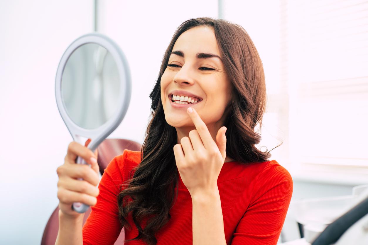 Hilarious girl with long curly hair in the dental chair of tooth center is using a mirror to see the result of the stomatology work which was done perfectly.