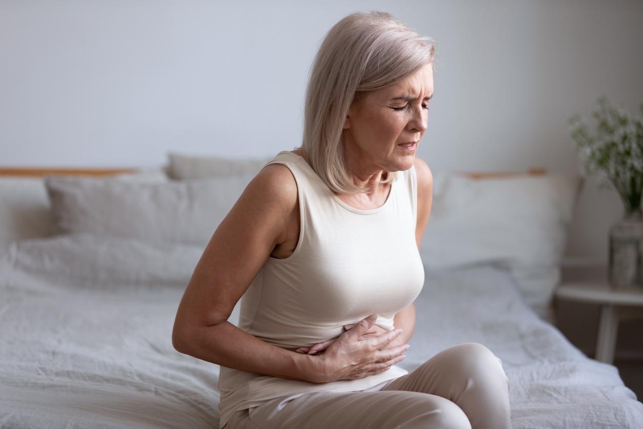 Unhealthy middle aged frowning woman sitting on bed, putting hands on belly, suffering from strong stomach ache. Unhappy older lady having painful feeling in abdomen, pancreatitis gastritis symptom.