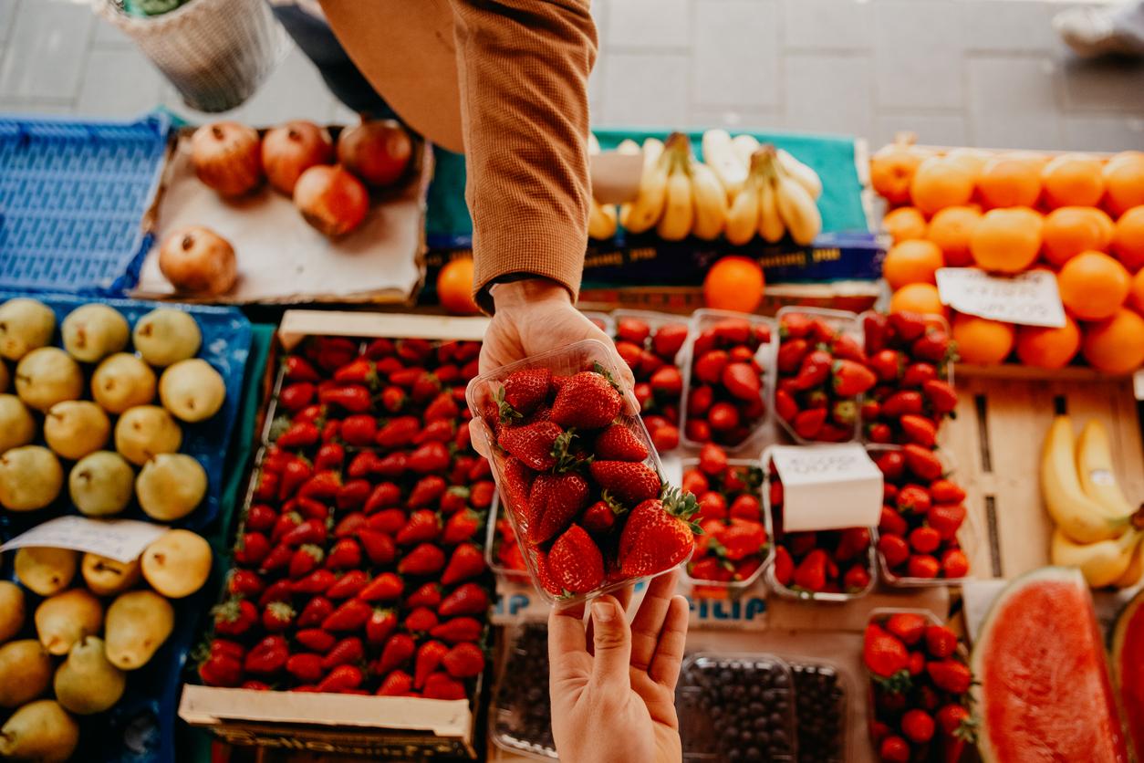 An app view photo of a male hand giving A box of fresh strawberries to a salesman hand, while the rest of the fresh fruits is looking great on the market table 