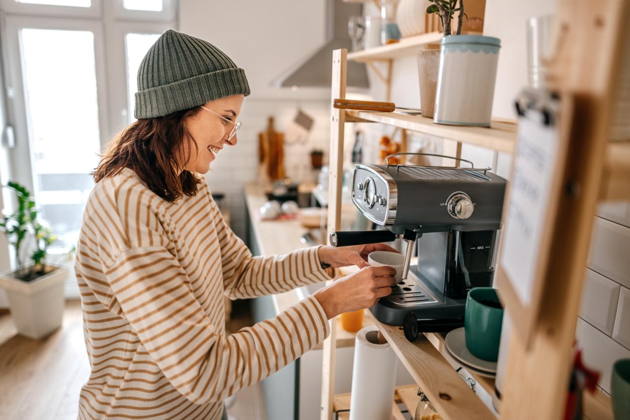 Young woman wearing knit hat preparing coffee through machine while standing in kitchen at home