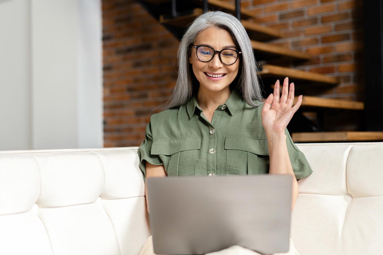 Mature female looks at webcam and greeting colleagues, waving and smiles sitting on sofa at home. Happy senior woman using laptop computer for video connection from home. Video call concept