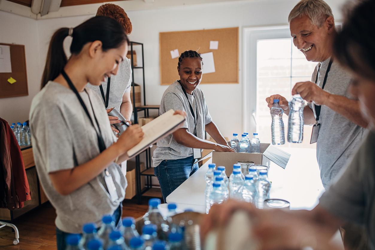 Diverse group of male and female volunteers sorting donated canned food and water bottles into cardboard boxes in charity center.