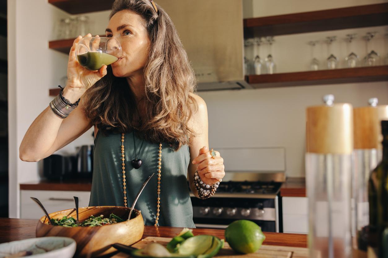 Vegan woman drinking some green juice while having a buddha bowl. Mature woman serving herself a healthy plant-based meal in her kitchen. Senior woman eating clean at home.