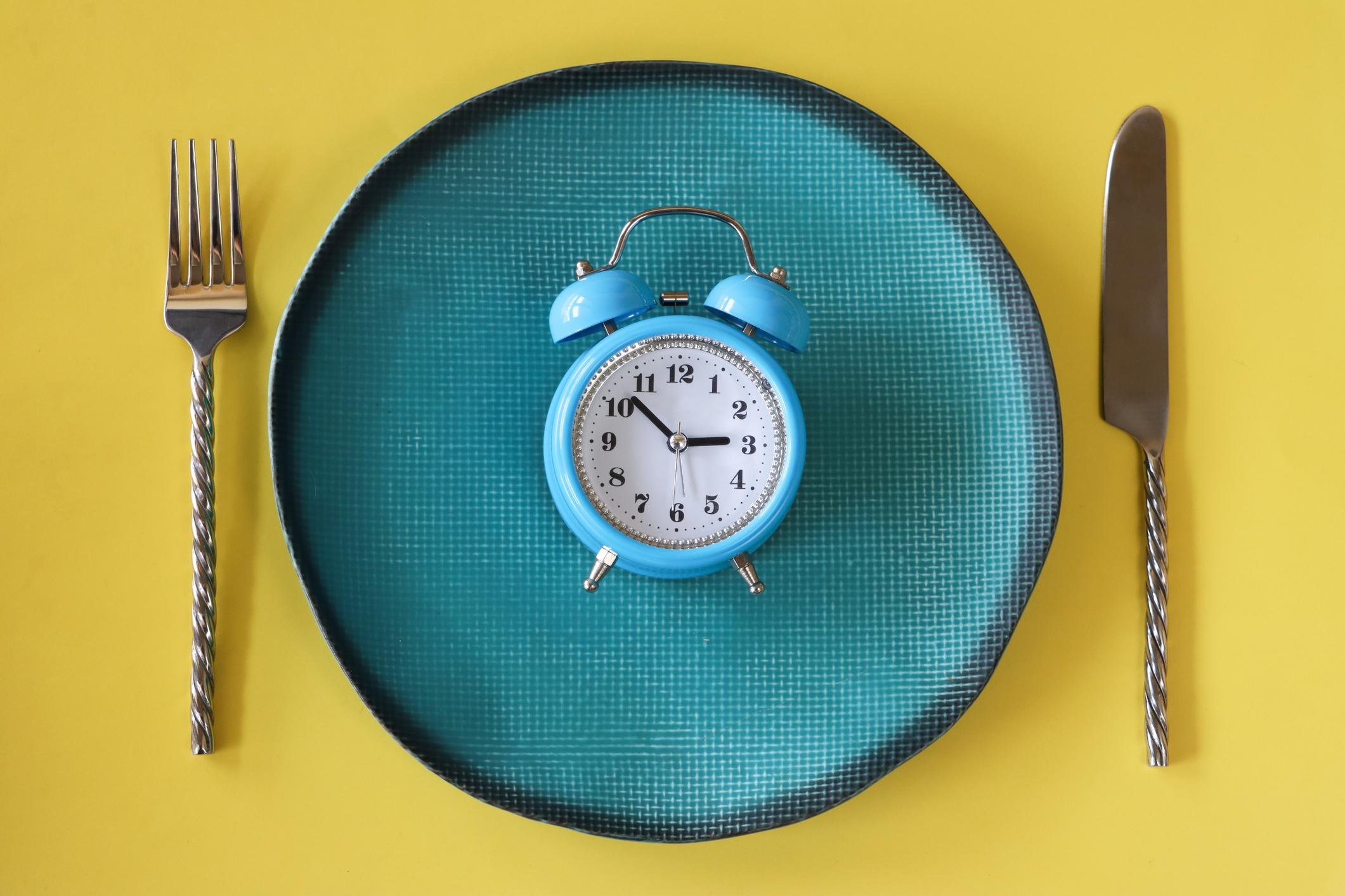 Stock photo showing close-up, elevated view of a healthy eating and intermittent fasting diet concept depicted by a plate containing a double bell alarm clock.