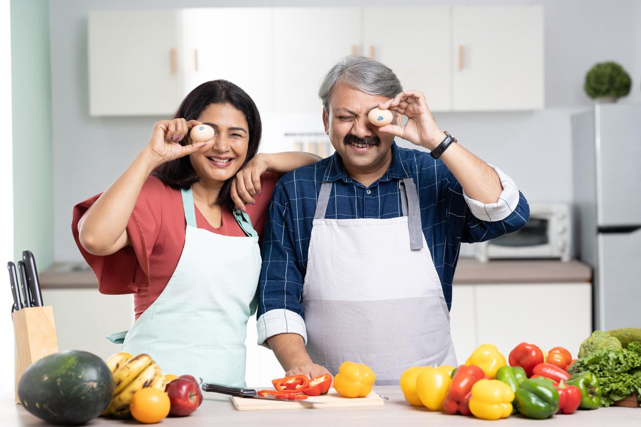 Indian couple in the kitchen holding eggs up to their eyes and preparing vegetables
