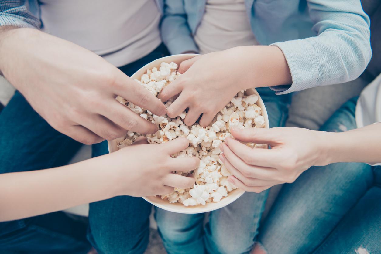 Top view close up portrait of hands taking sweet pop corn from bucket, dad mom kids sharing popcorn while watching film program tv, sitting on sofa indoor, enjoying free time having fun