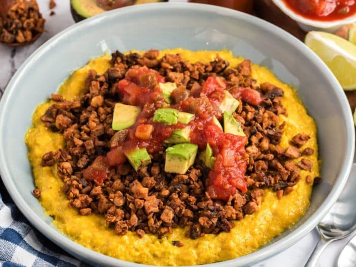 a vegetarian or vegan dish featuring a bed of creamy yellow polenta or similar grain topped with seasoned crumbled soy-based meat substitute (likely “ground” tvp), diced avocado, and salsa