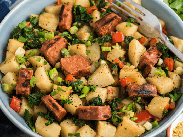 a close-up of a bowl of potato hash. The hash includes diced potatoes, pieces of what appears to be grilled sausage or a sausage substitute, chopped kale, green onions, and red bell peppers